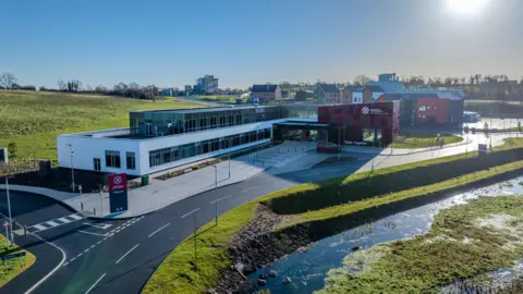 Training centre drone image, the main building has a white and red painted front, with NIFRS and the service emblem on the red wall. It sits by a water feature and grass. 