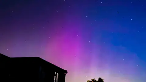 BBC Weather Watchers/UkWeatherView Above the roof of a house, which is a dark silhouette, the sky is bright purple into dark blue with lots of very bright stars