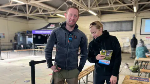 A man and woman are standing together. The man, on the left, is looking at the camera and holding a dog lead. The woman, on the right, is looking to the floor. 