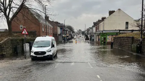 A white Northumbrian Water van parked on the side of the flooded street. A section of the road, and the surrounding pavement, is covered in water. A passer-by is standing outside Sultan Halal Superstore and looking at the flood.