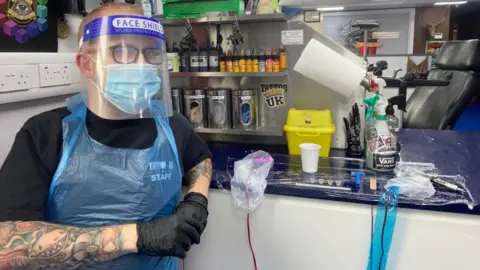 ADAM GRANT Tattoo artist Adam Grant wears a face mask, visor and disposable gloves and apron.