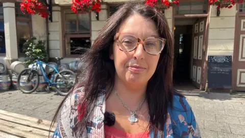 A woman wearing an orange top under a colorful jacket and glasses stands outside a pub while looking into the camera.