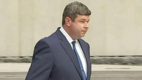 RTÉ Shane Flanagan in a grey suit and blue tie pictured leaving court. 
He has short black hair and his hands are in his pockets. 
