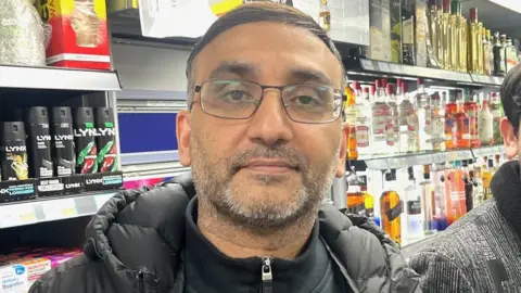 A mna with a grey beard and glasses wearing a black parka jacket and zipper. He is standing in front of a shelf full of lynx deodorant and alcohol.