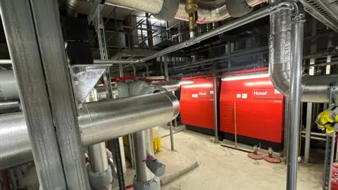 Two red commercial gas boilers are surrounded by a series of insulated pipework which is silver in colour. There is white writing on the boilers with the word Hoval.