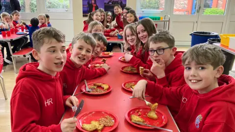 A group of children sit at a dinner table enjoying their school meals. 