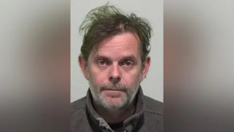 Mugshot of William Henderson who messily ruffled dark hair and a grey beard.
