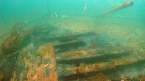 Bournemouth University An underwater photo of a shipwreck. Timbers can be seen with algae growing on them. The water is green/blue and the wreck site on the seabed is marked out. 