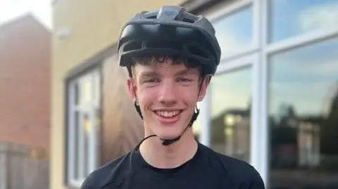 Sarah Sanderson/BBC A landscape head and shoulders shot of a teenager in a black top with a white tick on it wearing a black helmet. He is smiling into the camera in front of a house.