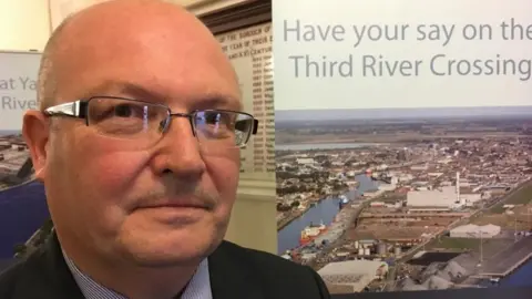 ANDREW TURNER/BBC Councillor Graham Plant next to Herring Bridge consultation sign