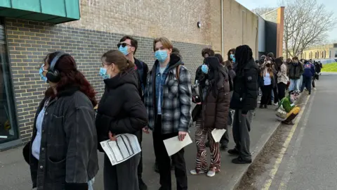Several people in jackets and blue facemasks, queuing along the side of a brick building.