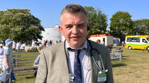 Shaun Whitmore/BBC David Martin, a man standing on a grassy field at the Royal Norfolk Show. He is looking directly at the camera and is wearing a shirt, blue tie and blazer.