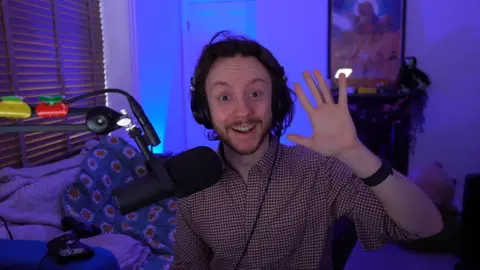 MrROFLWaffles A man smiles, waving at the camera. He's in a room set up for livestreaming, equipped with a microphone on a boom arm and with lighting that lends it a blue hue.