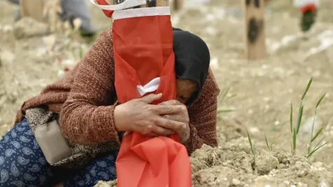 Goktay Koraltan/BBC Fethiye Keklik clutching the makeshift grave marker where her son, who died in the quake, is buried