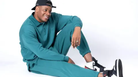 Tolu Tolu Ogunmefun sitting on the floor with his knee up, posing in a turquoise tracksuit and cap