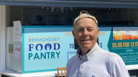 Ken Blower wearing a white t-shirt and a cap on backwards stands in front of a shop front that says food pantry