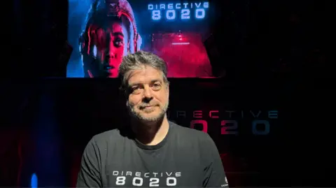 Danielle Malgwi The image shows a man wearing a black shirt that says "Directive 8020", with a key art sign behind him showing the same words.