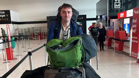 Joshua Pritchard is loaded up with luggage in an airport. He has a bag on his back and a pile of them on a trolley he is pushing. Behind him the airport is empty but for two people and three staff members at a check-in desk.
