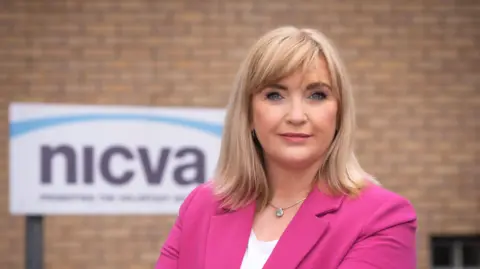 Celine is standing outside in front of a brick wall with a sign that says 'NICVA' behind her. She is wearing a bright pink blazer, white t shirt, silver necklace and she has blond hair to her shoulders. She has blue eyes and is wearing pink lipstick. 