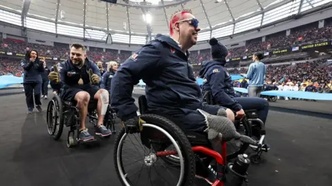 Reuters A man in a wheelchair and spiked hair dyed red in the foreground as other teammates in wheelchairs are behind him in the middle of the stadium