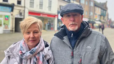 Elizabeth Baines / BBC A woman with short blonde hair wears a lilac waterproof coat. She stands next to a man with a grey beard, grey coat and grey flat cap