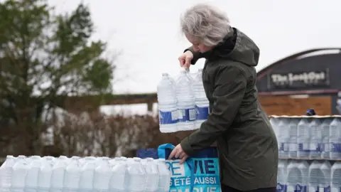 PA Media A woman places a case of bottled water into a shopping bag. The bag is held up on a large pile of water cases.