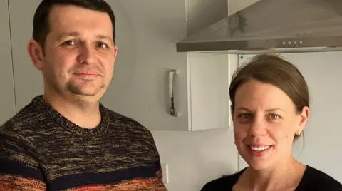 A man wearing a jumper is on the left next to a woman with dark hair wearing a dark top. They are in the kitchen looking at the camera. 