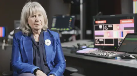 Baroness Helena Kennedy sits in a broadcast studio, wearing a bright blue blazer with a dark top and a white brooch. She has a pensive expression and behind her, several monitors glow with colourful test patterns and control screens, surrounded by keyboards and technical equipment.