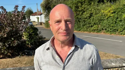 Luke Deal/BBC A bald man stands beside a road in a village. He is smiling and is wearing a light blue/grey shirt that is slightly open around his chest. There are buildings and bushes in the background. 