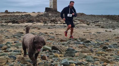 Philip's Footprints A man in a black athletic outfit with the number '2' runs along a rocky beach, following a brown curly-haired dog. In the background, an old stone tower stands on a small rocky hill under an overcast sky