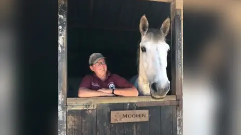 Wiltshire Police Jayne Hunt standing next to a white horse. She is wearing glasses, a hat and a burgundy polo shirt. Her arms are on top of a stable door and she is looking at the horse.