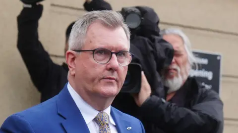 Jeffrey Donaldson leaving court during his trial. He has short grey hair and round-rimmed glasses. He is wearing a blue suit with a white shirt and a blue and gold tie. Behind him a man with white hair and beard can be seen holding up a broadcast TV camera filming him. He is wearing a dark raincoat.