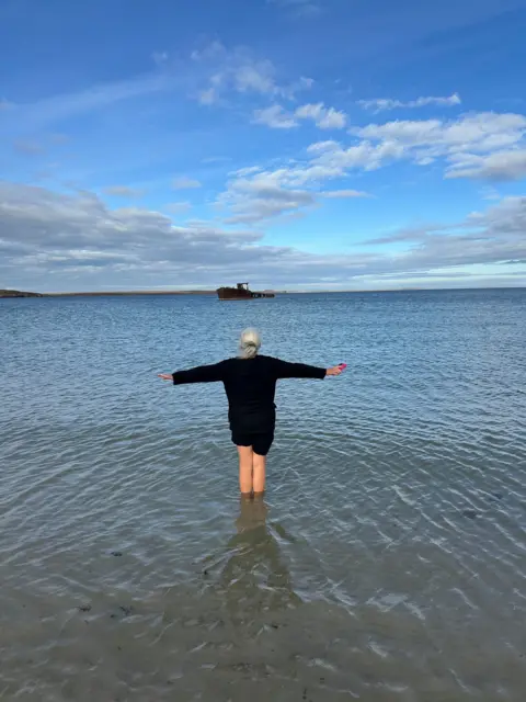 Derek Gourlay Woman standing in water with her back to the camera and arms outstretched, looking out over the water with a rusted shipwreck in the far distance