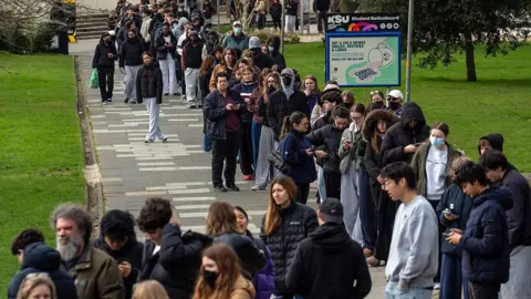 Getty Images A long queue of young people stretches along a paved path on a university campus, many wearing dark coats and some wearing face masks. A Kent Student Union noticeboard and a Campus Coffee sign are visible beside the path. Green grass and trees line the walkway in the background.