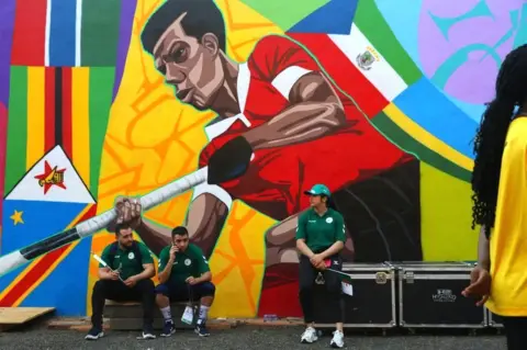 NIPAH DENNIS/AFP People in Algeria kit sit on the sidelines next to a large mural.