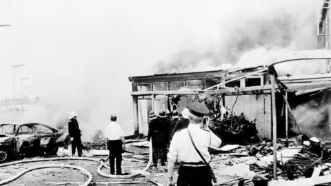 PA Media A dark mushroom cloud of smoke drifts across the centre of Belfast in July 1972, as firemen hose down the remains of Oxford Street bus station
