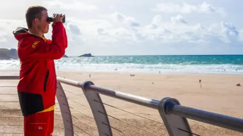The image shows a man standing on a raised seaside viewing platform, looking out over a wide sandy beach and the sea. He is wearing a bright red jacket and red shorts with black and yellow detailing. The letters RNLI are visible on the sleeve of the jacket, along with Lifeguard text on the shorts.