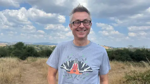A man with a butterfly logo T shirt in a field with hills behind.
