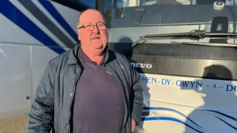 A man stood in front of a bus. He is wearing a blue coat and a blue jumper. He has glasses, and a moustache.