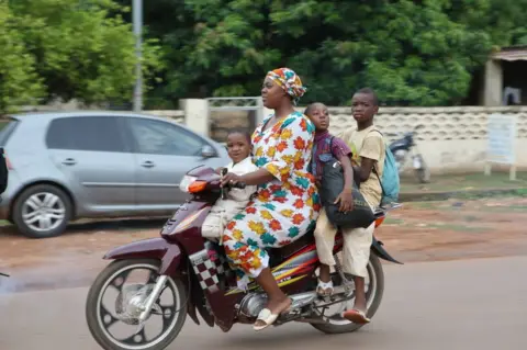 Getty Images SIKASSO, MALI - JUNE 25: Malian woman rides a motorcycle with her children as daily life amid poverty continues in West African country Mali's Sikasso on June 25, 2022