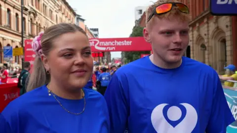 A young woman and young man are smiling as they stand next to each other. Both are wearing blue The Christie Charity T-shirts.