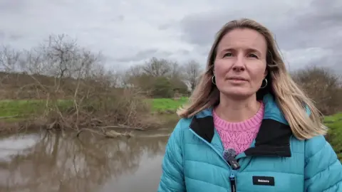 Aimee Felus from Western Sussex River Trust stood next to a river. She is wearing a blue puffy jacket and a pink jumper talking to the camera.