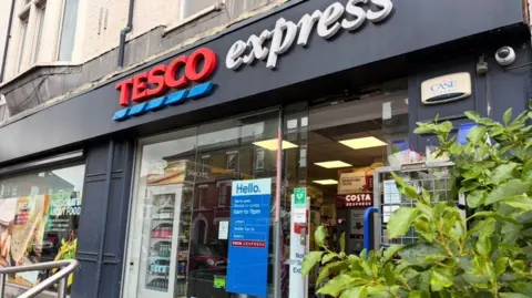 The exterior of a Tesco Express store doorway with green foliage next to it. 