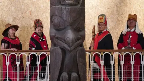 PA Media Earl Stephen's, second from left (who goes by the Nisga'a cultural name, Ni'is Chief Joohl) with Pamela Brown (left) as they join delegates from the Nisga'a Nation next to the memorial pole of 11 meters high during a visit to the National Museum of Scotland in Edinburgh, before the 11 meter high memorial pole returned to what is now British Columbia.