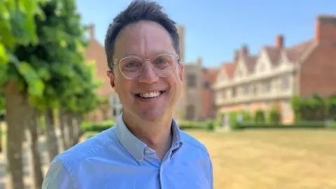 Oliver Burke, a man wearing glasses and a light blue shirt, is stood in front of an English Tudor building. There is a lawn behind him and a row of trees.