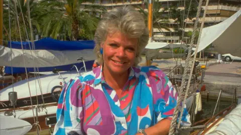 Getty Images Judith Chalmers in a bright multi-coloured blouse in front of a Mediterranean harbour on Wish You Were Here?
