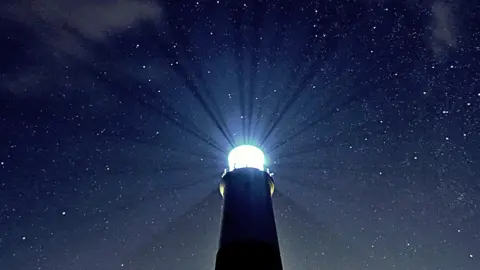 The sky is pitch black but studded with stars. In the centre stands the top of a lighthouse with its light shining bright. You can even see beams of light being cast from the lighthouse into the night sky.