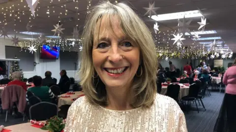 Kate Bradbrook/BBC Woman with long blond hair wearing sparkly top in a room full of Christmas decorations