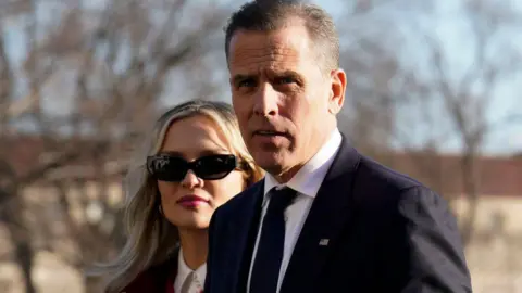 Hunter Biden in dark suit with dark tie with wife Melissa Cohen Biden who is wearing sunglasses in front of bare tree branches