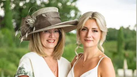 Simon Buck Christine Cullen and Emma-Jayne Herbert on Emma-Jayne's wedding day. Christine is wearing a designer brown wedding hat and glasses, in a cream short-sleeved dress, and is smiling at the camera. Emma-Jayne is wearing a thin-strapped white wedding dress, she has blonde shoulder-length hair tied back and is smiling at the camera